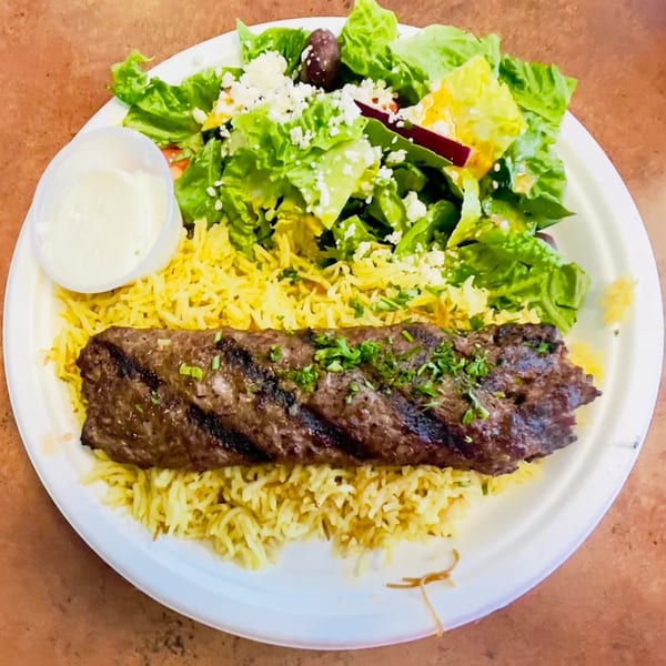 A Greek platter with grilled meat, yellow rice, a side salad with feta, and a cup of sauce.