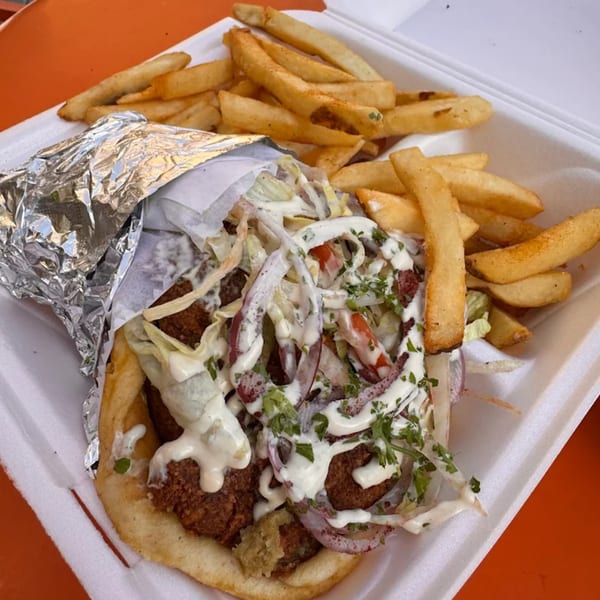 A Greek falafel pita with white sauce, red onions, and fries in a white takeout container.