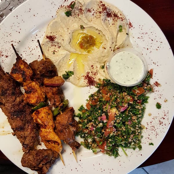 A Greek platter with grilled meat skewers, hummus, tabbouleh salad, and a side of tzatziki sauce.